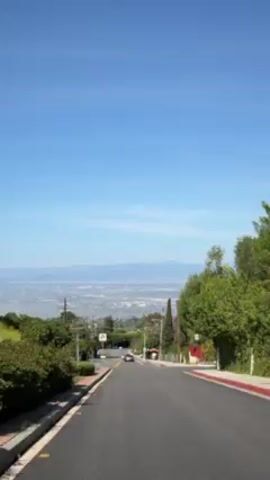 The video shows a road leading down a hill under a clear blue sky.  A car is seen driving down the road. On either side of the road are trees and houses. In the distance, a city is visible, backed by mountains.  There are utility poles and street signs along the road.