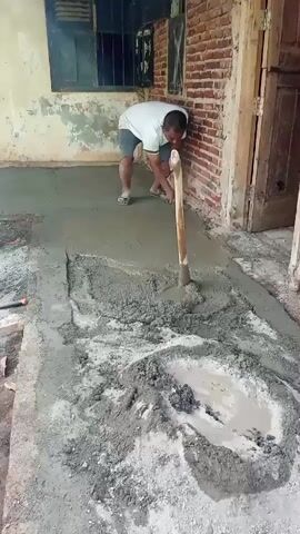 A man kneels on a concrete surface, using a wooden-handled tool to spread wet cement. He is wearing a white t-shirt and grey shorts. A pile of wet cement is in the foreground, with water pooling in the center. The man is working next to a brick wall, and there is a window to his left. The area appears to be an outdoor construction site.