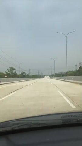 A first-person perspective video shows a vehicle traveling on a wide, paved highway under an overcast sky. The highway has multiple lanes separated by white lines. Tall streetlights line the right side of the road, and electrical pylons are visible in the distance to the left. Trees and some low buildings can be seen beyond the highway's barrier on either side. The road ahead appears to curve slightly upward into the distance. The dashboard of the vehicle is visible at the bottom of the frame.