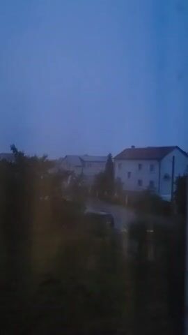 This media file captures a scene in Belaazyorsk, Belarus, during what appears to be twilight or a very overcast day. The dominant colors are deep blues and grays, suggesting a muted or dusky atmosphere. In the foreground, dark, blurred foliage obscures the lower part of the image, creating a sense of looking through a window or from behind a screen of plants.

The middle ground features several buildings. The most prominent is a light-colored, multi-story house with a reddish-brown roof. It has visible windows, and a white pole or pipe extends vertically along its side. To the left of this house, another building with a lighter gray roof is partially visible. A dark-colored vehicle is parked on a wet or reflective surface, possibly a road or driveway, in front of these structures. Trees and shrubs are scattered throughout the scene, adding to the impression of a residential or semi-rural setting.

The sky is a uniform, deep blue, devoid of clouds or any discernable celestial bodies. The overall lighting is dim, indicating either early morning or late evening. The wet appearance of the ground and the lack of sharp details suggest either rain has recently fallen or the atmosphere is very humid, contributing to a somewhat hazy or misty quality. No people are visible, and no text is present in the media. The mood evoked is calm, quiet, and perhaps a bit melancholic due to the dim lighting and lack of activity.