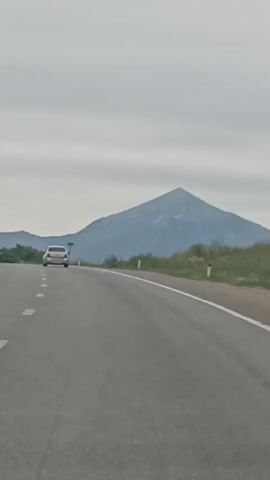 A white car travels on an asphalt road in a rural area. The road has white dashed lines marking the lanes and white posts along the side. In the background, a large, conical mountain with a rocky peak stands under a cloudy sky. The surrounding landscape is covered in green grass and some scattered bushes. The car is positioned on the left side of the road, moving away from the viewer. The shot is taken from a moving vehicle, indicating the perspective of someone in the same direction of travel as the white car.