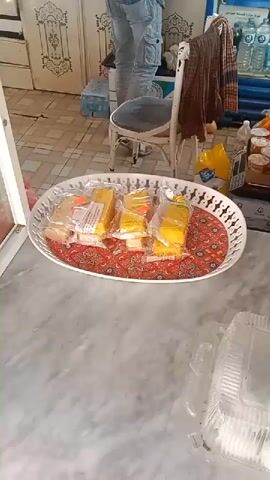 A man wearing a plaid shirt, blue jeans, and a white cap is standing in a room that appears to be a store or a kitchen. He is looking into a refrigerator and reaching for something. In the foreground, there is a white oval tray filled with individually wrapped yellow food items, possibly cakes or pastries, arranged on a patterned red cloth. The room has tiled floors and walls, with cabinets and shelves visible in the background. There is also a chair with a brown garment draped over it. The overall environment suggests a commercial or domestic setting where food is being stored or sold.