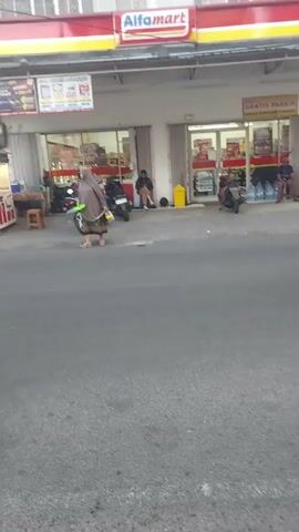 A dark-colored SUV drives past a street-level view of an Alfamart store entrance. A motorcycle with a rider is visible on the left, and a woman wearing a hijab walks away from the store towards the center of the frame, carrying a green bag. Several motorcycles are parked in front of the store.