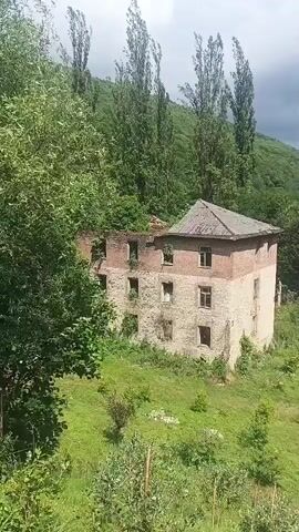 A drone view slowly pans across a dilapidated, multi-story brick and stone building surrounded by lush greenery and tall trees. The building appears abandoned and is in a state of disrepair, with some windows missing and vegetation growing on its exterior. The surrounding landscape is hilly and densely forested, with a cloudy sky overhead. The drone movement is smooth and continuous, revealing more of the natural environment as it moves.