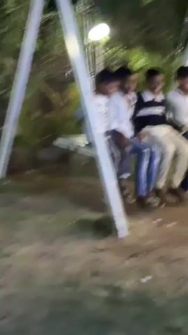 The video shows a small group of children outdoors. Initially, a child in a white patterned shirt and light-colored pants seems to playfully push at a person in a maroon shirt and jeans near a low wall. Then the scene shifts to a wooden swing set, with three boys sitting and swinging together. A fourth boy jumps on, and they all swing together. They are smiling and appear to be having fun. The surroundings suggest it is nighttime, as indicated by overhead lights, and there are trees and vegetation visible in the background.
