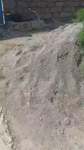 The video appears to be filmed on a sunny day, possibly in the morning or afternoon, in an outdoor setting that resembles a rural or industrial area. The ground is covered in dirt and gravel, with a large pile of what looks like sand or construction material in the foreground. In the background, there is a wall constructed of large stones, possibly part of a building or enclosure.

To the left of the frame, there is a partially visible structure that could be a well or a water tank, with a white pipe extending from it. Further to the right, a bright blue gate or door is visible, slightly ajar. Next to the gate, there are some green plants and a yellow bucket. A pile of black plastic bags or debris is also present near the wall and gate.

The camera pans slowly across the scene, offering glimpses of the surrounding area. The overall impression is one of a working area or storage yard, possibly at a construction site or a farm. There are no people visible in the video, and the activity taking place seems to be a static observation of the environment. The sun casts distinct shadows, indicating clear weather. The location context provided (Sisavan, Armenia) suggests this is an outdoor scene in that region. There is no visible text in the video.
