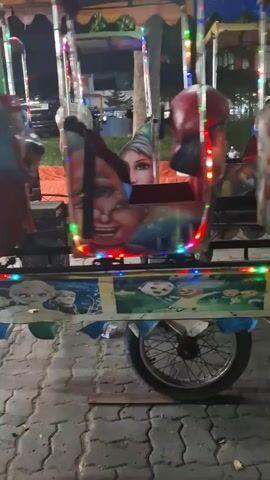 The video shows a close-up of a children's amusement ride at night. The ride consists of multiple colorful carriages, each decorated with cartoon characters, and is illuminated by strings of colored lights. The carriages appear to be in motion, as indicated by the slight blur. In the background, trees and some structures are visible, suggesting an outdoor setting. The ground is paved with interlocking stones, and a wheel of the ride, possibly attached to a motorcycle-like base, is prominent in the foreground. The overall atmosphere is festive and geared towards children.