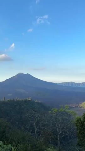 This is a vertical video of a mountain landscape. The video starts with a wide shot of a mountain range under a clear blue sky with a few white clouds. In the foreground, there are trees and lush greenery. The camera then pans slowly to the right, revealing more of the mountains and a body of water, likely a lake, nestled between them. The sun appears to be low in the sky, casting a warm light on the peaks and illuminating the mist that hovers around them. The overall impression is one of natural beauty and tranquility.