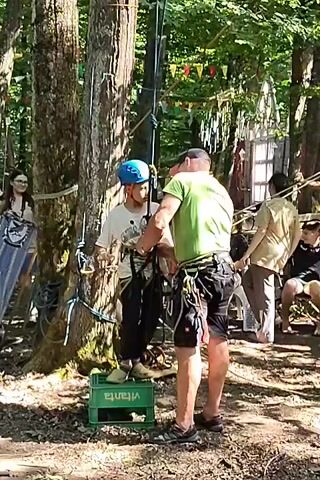 A group of people are in a wooded area, seemingly at an adventure park. In the foreground, a man in a green shirt and black shorts, wearing a cap and a harness, is assisting a younger person in a blue helmet and white t-shirt, who is also wearing a harness. They are standing near a tall pole and some ropes, with the younger person standing on a green crate. Other people are visible in the background, some standing and some sitting. There are trees, ropes, and what appears to be a small wooden structure. The overall atmosphere suggests an outdoor recreational activity, possibly zip-lining or a ropes course.