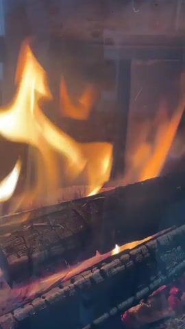 A close-up shot shows burning logs inside a fireplace. Flames are visible in various shades of orange and yellow, flickering and dancing around the logs. The logs themselves appear charred and are glowing red from the heat. The overall environment suggests a cozy and warm setting, possibly a home during colder weather. The camera is focused on the fire, with no people visible.
