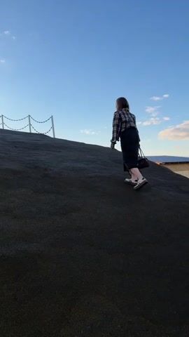 A woman in a plaid shirt and dark skirt walks uphill on a large, smooth, dark-colored rock or structure. There is a rope railing at the top of the hill. As she nears the top, two other people become visible: one sitting on a chair and another standing. The sky is blue with some clouds.