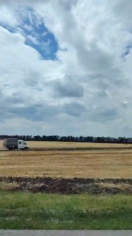 This video captures a view from a moving vehicle, likely a car, traveling through a rural landscape in Kagal'nitskaya, Russia. The scene is dominated by vast fields of golden, dry grass or wheat, suggesting a post-harvest period or a dry climate. The sky is partly cloudy with dramatic, billowy white and gray clouds, indicating a potentially changeable weather system, possibly during daytime given the brightness.

In the distance, a line of dark trees borders the fields. A white truck with a covered bed is seen driving on a dirt path or road within the field, kicking up dust. The video transitions to show road markers with black and white stripes along the side of a gravel road in the foreground, with the fields and trees continuing to stretch into the distance.

Later, the view shifts to looking out the car's windshield, revealing a paved road with white lane markings. Another white vehicle is visible driving ahead on this road. The interior of the car is partially visible, including a dashboard with air vents and part of the car's window frame. The overall impression is one of a journey through an agricultural region under a dynamic sky. No text is visible in the video.