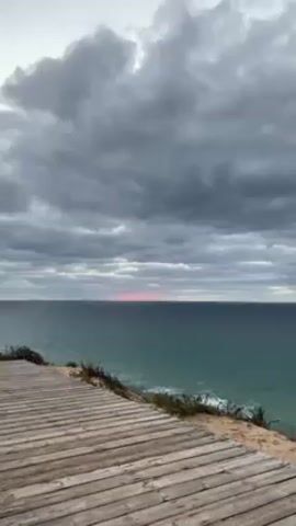 The video shows a body of water, likely the ocean, under a sky full of gray clouds. A sliver of pinkish-red color appears along the horizon line where the sky meets the water. In the foreground, there is a wooden boardwalk or viewing platform, suggesting that the viewpoint is from an elevated coastal area. Some vegetation can be seen on the side of the platform. No people are visible in the video.