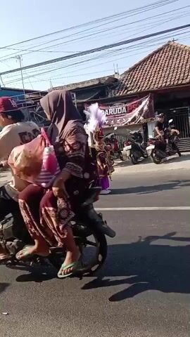 A parade is taking place on a street. There are several people in elaborate costumes. One person is wearing a tall, ornate headdress with purple feathers and a long, flowing purple skirt. Another person is dressed in a detailed gold and black costume with a large headdress. There are also people in more casual clothing, some watching the parade and others participating. Motorcycles are parked along the side of the street and one is being ridden. A banner with the word "GARUDA" is visible on a building in the background. The scene appears to be in a sunny, urban or suburban environment.