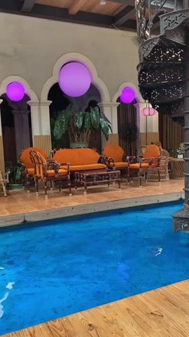 A bird's eye view shows a blue pool with a wooden deck. A spiral staircase is visible to the right. Orange furniture, including chairs and a couch, is arranged on the deck. Large purple orbs are suspended from the ceiling. Lush green plants are placed throughout the space. A pink structure with a thatched roof is visible in the background. The overall atmosphere is a stylish indoor/outdoor lounge area.