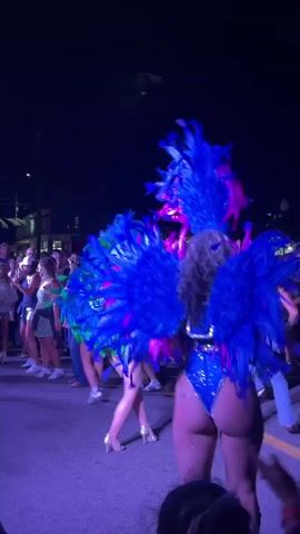 The video shows a street performance at night, illuminated by purple and blue lights. Several people in elaborate, feathered costumes are dancing in the foreground, with a crowd of onlookers lining the street behind them. The costumes feature large blue and green feathers, sequins, and headdresses. The dancers are wearing high heels and short, revealing outfits. The crowd appears to be enjoying the performance, with some people taking pictures and videos. The overall atmosphere is festive and energetic.