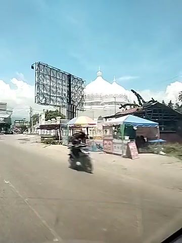 A car is driving along a street toward Masjid Al-Istiqomah. The mosque is white and features two large domes. A large billboard is situated near the mosque. A motorcycle drives on the street past the car. The sky is blue with some white clouds. There are other buildings, trees, and power lines visible. Some small structures with canopies are situated alongside the street.