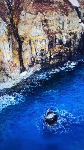This media file is a short, still video clip showcasing a dramatic coastal landscape. The scene is dominated by a rugged, sheer cliff face that slopes down to meet a turbulent, deep blue sea. The cliff is textured with layers of rock in various shades of brown, tan, and grey, with patches of snow clinging to its upper reaches and a thin streak of water cascading down one side.

At the base of the cliff, a rocky shoreline meets the water, where waves crash and foam, creating a dynamic contrast with the stillness of the cliffs above. A single, dark, rounded rock juts out of the water in the foreground, surrounded by swirling whitecaps.

The overall impression is one of a wild, untamed natural environment. The lighting suggests it is daytime, with clear, bright conditions that illuminate the colors of the rock and the intensity of the blue water. There are no people or man-made objects visible, emphasizing the raw beauty of the location. The lack of specific landmarks makes it difficult to pinpoint the exact location without additional context, but the stark, rugged coastline is a common feature in many parts of the world. The media file does not contain any visible text.