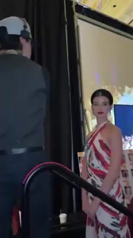 The video shows a fashion show backstage. Several models are lined up, wearing dresses in reds, whites, and floral prints. One model is in a white dress with red and grey brush strokes. Another is in a red dress with floral prints. There is a large screen displaying the runway portion of the show. Staff members stand nearby, presumably helping the models. The room has high ceilings and patterned walls. People are present, watching the models and the show on the screen.