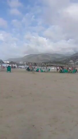 A wide shot of a sandy beach with scattered people and umbrellas. In the background, there are hills covered in vegetation and buildings. The sky is cloudy. The camera pans slowly from left to right.