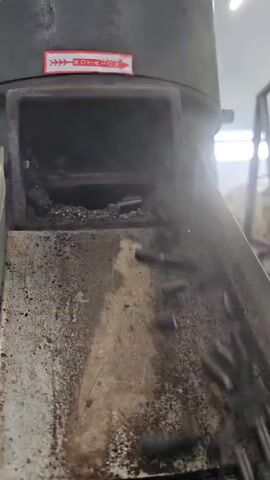 A close-up shot shows charcoal pellets falling from an industrial machine. The pellets are dark gray and cylindrical, approximately one inch in length and a quarter inch in diameter. They are dropping from an opening at the top of the machine into a funnel-like chute below. The chute is stained with residue, suggesting it has been used for processing similar materials. At the top of the machine, a red and white sticker with arrows and the word "ROTATION" is visible, indicating the direction of movement of an internal component. Steam or smoke emanates from the opening where the pellets are falling, suggesting a process involving heat or combustion. The background is blurred but appears to be an industrial setting, with some indeterminate objects and lighting. The video focuses on the continuous production of charcoal pellets.