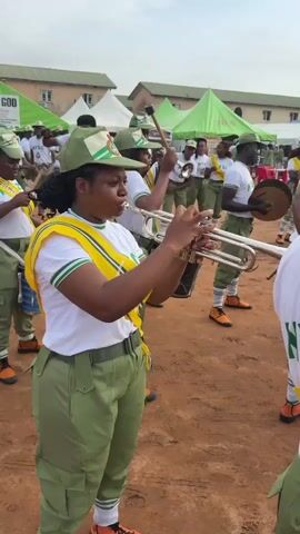 This media file features a group of young people dressed in matching uniforms, participating in a band performance. The subjects are primarily individuals playing musical instruments, including trumpets and drums. They are wearing green cargo pants, white t-shirts with yellow sashes, and green caps with a circular emblem. Their orange shoes stand out against the dirt ground.

The scene is set outdoors, with buildings and green tents visible in the background. One tent has visible text that reads "ABLE GOD". The overall atmosphere appears to be a daytime event, with overcast or diffused lighting suggesting cloudy weather. The participants seem focused on their performance, with some smiling and others concentrating on playing their instruments. The event appears to be an organized gathering, possibly a parade or a ceremonial band performance, given the uniformed participants and musical instruments. The location is identified as Alagba, Nigeria.