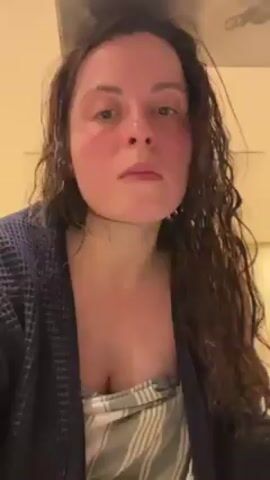 A woman with long, curly brown hair stands in front of a camera, wearing a blue bathrobe. She looks directly at the camera, and after a moment, she brings her hands up to her face, holding some white material. She blows on the material and moves her hands away from her face. She then touches her neck and rubs her forehead with her hands. The background is a blurry light yellow wall.