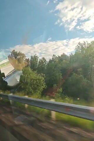 The video is a first-person perspective from inside a moving vehicle, looking out the side window at a passing rural landscape. The sky is blue with scattered white clouds, and the sun appears to be somewhat low, casting a warm glow. Trees and green vegetation line the side of the road, and in the background, a few houses with varying roof styles can be seen. A yellow diamond-shaped road sign with a white border is visible near the beginning of the clip, and what appears to be a power pole passes by shortly after. The reflection of the vehicle's interior, possibly including part of the dashboard or seats, is visible in the window throughout the video. The view is dynamic as the vehicle moves forward.