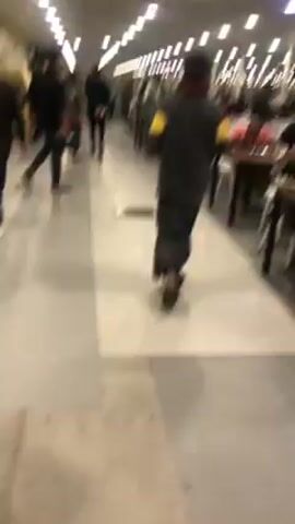 The video shows a brightly lit hallway in a building. The floor is a light-colored tile with linear, darker inlays. Numerous individuals, mostly men, are walking along the hallway in different directions. Many are wearing dark clothing, with a few exceptions, such as someone in a light-colored garment and another person with yellow armbands. Some tables and chairs are visible along one side of the hallway. The camera is moving, presumably being carried by someone walking, and the view is slightly shaky.