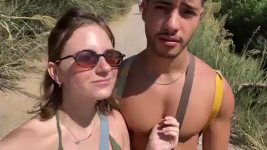 A young couple is walking along a dirt path. The woman wears sunglasses and a green and blue tank top, and the man is shirtless with a yellow strap over his shoulder. They are surrounded by tall, dry grass. The man looks at the camera and smiles, then kisses the woman on the cheek. The sky is visible in the background.