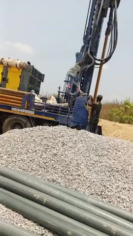 The scene shows a blue and yellow water well drilling truck in an outdoor setting. A man is seen standing on the truck bed near a large yellow generator that is emitting black smoke. Another man stands near the drilling rig.  A large pile of gravel is in the foreground, along with several gray pipes. The sky is mostly clear with some scattered clouds. The phone number 08184955555 is visible on the side of the truck.