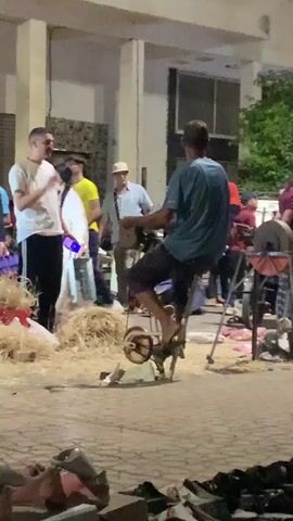 The video shows a man sitting on a stationary bike-like device in an outdoor setting. The device appears to be manually powered and connected to a machine that seems to be used for grinding or sharpening. The man is dressed in a blue shirt and dark shorts, and he is pedaling the device with his bare feet. There are several other people around him, some standing and watching, others walking by. In the background, there is a building with columns and a window. There is also a pile of hay and some scattered debris on the ground. Some individuals appear to be wearing headscarves.  The lighting suggests it is dusk or night.