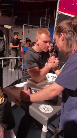 The video shows an arm-wrestling match between two men. The man on the left has short red hair and is wearing a black t-shirt, while the man on the right has long brown hair in a bun and is wearing a blue t-shirt. They are seated at a table with stickers on it, facing each other with their elbows on the table and hands clasped, ready to start. A few other people are observing the match in the background, including a woman with glasses and a man in a black t-shirt standing by some equipment. There is a sign in the background in Russian. The environment appears to be a sporting event.