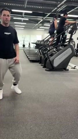 A man in a black t-shirt and grey sweatpants walks in a gym. He passes by a row of stair climbers where two women are exercising. He then walks towards a cable crossover machine. He stops and bends down to pick up a black attachment from the floor. He stands up and looks at the camera.