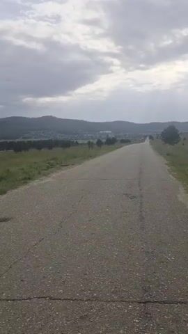 The video shows a paved road stretching into the distance, bordered by grassy fields and a line of trees. In the background, a town or village is nestled at the foot of rolling hills under a cloudy sky. The road surface appears worn with cracks. The camera is moving forward along the road.