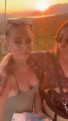 The video shows three young women in a cable car during sunset. The woman in the foreground is on the left, wearing a green top and sunglasses on her head. She is smiling and looking at the camera. The woman in the middle is wearing a patterned top and sunglasses, and she is making a duck face. The woman on the right is wearing a yellow top and is looking out the window. The background shows mountains and a town, illuminated by the setting sun. The women are holding a bottle of water, and there are other bags and personal items visible in the cable car.
