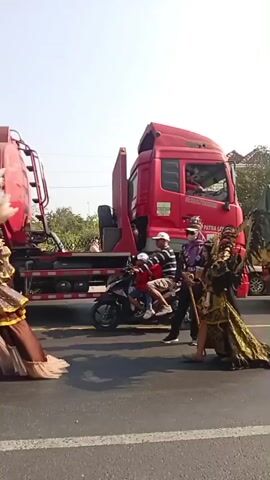 The video shows a street scene with several people in elaborate costumes walking in a procession. In the foreground, a person on a motorcycle is visible, partially obscuring the view. Behind the motorcycle, a large red truck is parked. To the right of the truck, several individuals dressed in ornate, colorful costumes are walking. One costume appears to be gold and black with a long train. Another features a peacock-like headdress and bright colors. Other individuals in various costumes and regular clothing are also present. A green-covered truck is parked further in the background. The sky is bright and clear, suggesting daylight. The procession is happening on a road, indicated by the presence of vehicles and the asphalt surface.