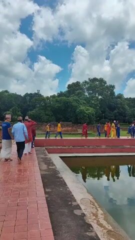 The video shows an outdoor scene with several people walking along a paved pathway. The pathway is made of reddish-brown bricks and has a railing on one side. To the right of the pathway is a body of water with reflections of the sky and trees. In the background, there are more people walking, some wearing colorful clothing. Beyond them, there are green trees and a blue sky with white clouds. Further in the distance, a large white building with windows and a flag on top is visible. There are also grassy areas and some brick structures scattered around the landscape. The overall impression is of a public place, possibly a park or a historical site, with people touring the area on a cloudy day.