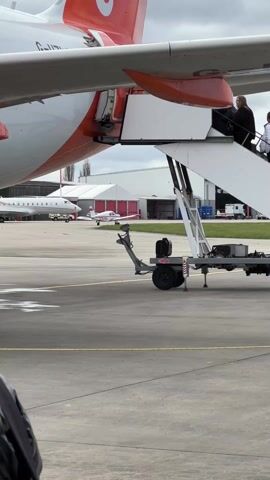 The video shows people disembarking from a white and orange plane using a set of portable stairs. The stairs are attached to a trailer with a single wheel. In the background, there are other airplanes parked near a hangar, and a white SUV is driving by. The day appears overcast.