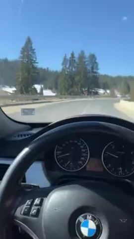 The video shows the view from inside a car driving on a road on a sunny day. The road is bordered by a metal guardrail and a snow-covered landscape with trees. The camera pans to the left to show a Bulgarian flag waving in the wind next to the road. The interior of the car is visible, including the dashboard, steering wheel, and side mirror. The sky is clear and blue.