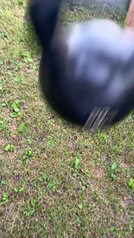 A hand grips a black kettlebell with a visible "20" weight marking. The scene is outdoors with green grass and some small weeds. The camera moves rapidly, suggesting a walking or running motion while holding the kettlebell.