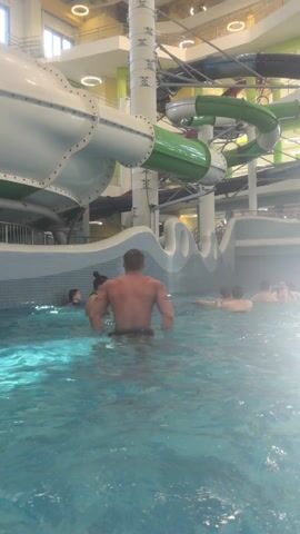 The video shows the interior of a water park, focusing on a man standing in a pool. He is facing away from the camera, and he is shirtless, showing a muscular back. There appear to be tattoos on his arms. Surrounding him in the pool are other people, including a woman with her hair in a bun, and other individuals further in the background. The pool water is clear with a light blue tint. The backdrop includes large water slides, some colored green and white, and a blue and white tiled wall designed with wave-like patterns. The overall lighting in the water park appears bright.