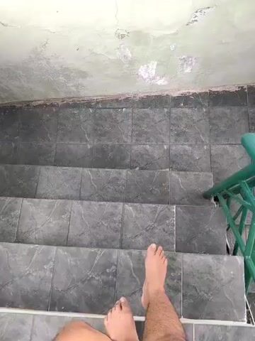 The video begins with a close-up shot of a person's bare feet and legs standing on a tiled surface. The camera then pans upwards, revealing a wall with peeling paint. The person takes a step down, and the camera continues to follow their movement down a set of stairs. The stairs are also tiled and appear to be old and worn. A green railing is visible on the right side of the stairs. The video ends with a shot looking down the stairs as the person continues to descend.