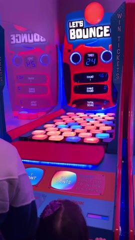 Two individuals, one with their arm and hand visible on the left and another with a hand visible on the right, are actively playing a brightly lit arcade game called "LET'S BOUNCE." The game is predominantly red and blue with glowing neon lights, set in what appears to be an indoor entertainment center.

The "LET'S BOUNCE" game features a main display showing a "TIMER" counting down (from 24 seconds at the start of the video), a current score (fluctuating, around 577-882 points), a "BONUS ROUND" score (0), and a "TOTAL SCORE" (0). Below this display is a grid of numerous square tiles that light up in various colors like orange, blue, and pink.

At the bottom of the game, there are two large, illuminated buttons labeled "1 POINT" and "100 POINTS." Between these, text on the game instructs players to "CLEAR ALL ORANGE TILES TO UNLOCK BONUS ROUND." Another button reads "MAKE GAME GO NOW." One player is seen holding and dropping a small orange ball onto the tile grid, attempting to score points. The adjacent game on the left is a mirrored reflection of the main game. The goal of the game seems to be to hit the tiles with the ball to accumulate points and tickets, indicated by the "WIN TICKETS" sign on the right side of the machine.