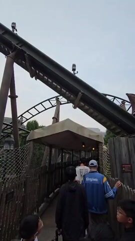 The video shows a roller coaster line at an amusement park. The roller coaster cars are going up an incline track. People are walking in a line towards a covered area. The area has wooden fences and netting. The sky is overcast.