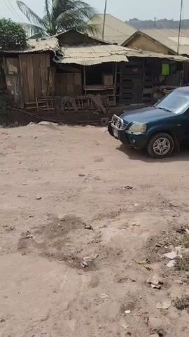 The video shows a dirt road in what appears to be a residential area with somewhat run-down structures. A green SUV is parked on the side of the road. A motorcycle with a driver and a passenger wearing a bright red puffy jacket passes the SUV. The SUV then drives off, and another motorcycle carrying two people drives down the same road. The environment looks dry and dusty. A palm tree is visible behind the buildings.