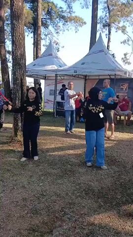 This is a video taken outdoors, likely in a park or campsite, with several people present. In the foreground, two women are facing the camera, performing some kind of dance or synchronized movement. The woman on the left is wearing a black shirt with yellow accents and black pants. The woman on the right is wearing a black hijab, a black shirt with yellow accents, and light blue wide-leg pants. Both have their arms extended to the sides. Behind them, a man in a gray t-shirt and blue jeans is also facing the camera and mirroring the women's arm movements. Further back, there are tents with logos, suggesting an event or gathering. Other people are visible in the background, some standing and some seated. The ground is covered in dry grass, and tall trees are present, with a clear sky overhead. The video captures a moment of group activity outdoors.