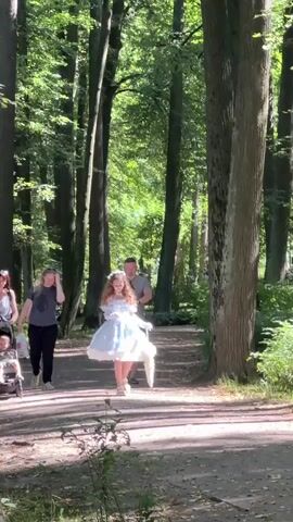 A young girl in a light blue and white dress walks along a path in a park, holding a white parasol. She has long, curly brown hair and is wearing white gloves. A man walks behind her, wearing a gray t-shirt and shorts. Sunlight filters through the trees, casting shadows on the path. Another person is partially visible in the background, pushing a stroller. The scene appears to be a leisurely walk in a wooded area.
