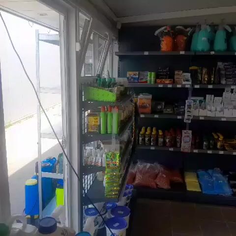 The video pans across the interior of a retail store that appears to specialize in agricultural or gardening supplies. On the left, a large window offers a view of the outdoors, where a blue barrel is visible. Inside, shelves are stocked with various products. The top shelves hold colorful spray bottles and containers. Below these, numerous smaller items are neatly arranged, including boxes, bottles, and packets. Some shelves display bulk items like bags of feed or fertilizer. Towards the right side of the store, more shelves are filled with rows of bottles and packets. The lighting appears to be artificial, coming from overhead fluorescent fixtures. The flooring is a dark tile or concrete. No people are visible in the video.