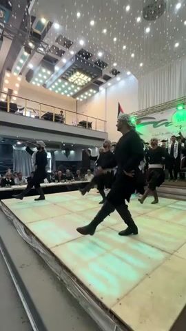 Four men in black clothing and head coverings perform a dance on a raised stage. The man in the foreground is wearing tall black boots and holding a curved sword. The other three men are also wearing dark clothing and appear to be holding similar items. The stage is lit with white and green lights. In the background, there is an audience seated at tables, a balcony area with people, and a banner with Arabic text and flags. The ceiling of the venue is decorated with numerous small, bright lights.